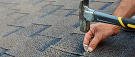 A roofer's hand hammering a nail to install new architectural roofing shingles on a home in Lansing, Michigan. A roofer's hand hammering a nail to install new architectural roofing shingles on a home in Lansing, Michigan.
